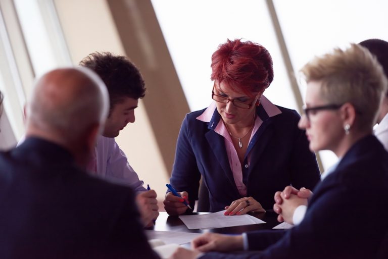 A group of people in business attire sit around a conference table. The focus is on a woman with short red hair and glasses, writing on a paper about key man insurance planning. Others are engaged in discussion, some partially visible. The setting appears professional, with natural light from windows.