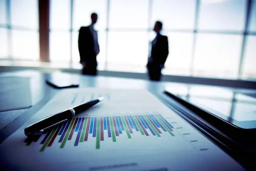 A close-up of a document with a color-coded bar graph and a pen resting on a desk. In the blurry background, two business professionals discussing key man insurance planning are engaged in conversation near large, bright windows.