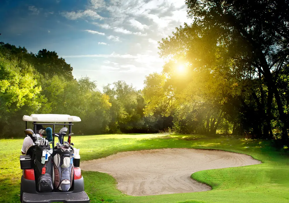 A golf cart filled with golf bags is parked next to a sand bunker on a sunny golf course. The green, tree-lined fairway stretches into the distance under a partly cloudy sky, with the sun shining brightly through the trees—a perfect setting to unwind and ponder retirement planning.