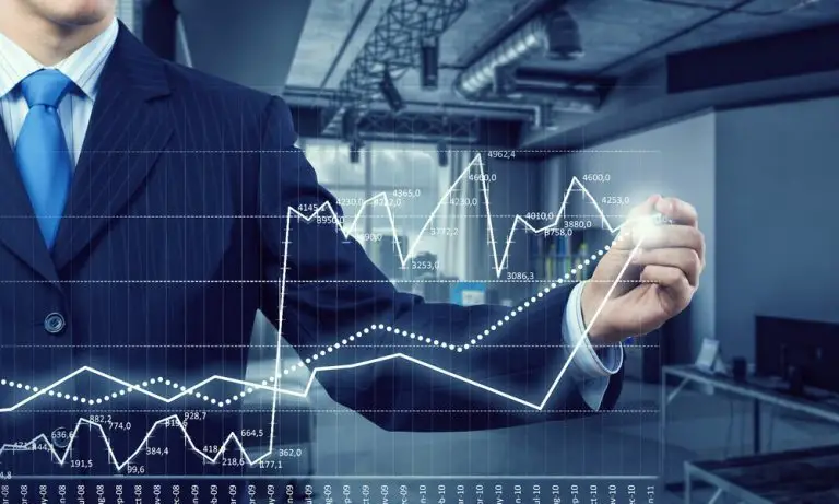 A person in a suit and blue tie stands in a modern office, using a digital pen to interact with a virtual financial graph displaying fluctuating data points and upward trends. The background features office furnishings and equipment, underscoring the importance of key man insurance for hedge funds.