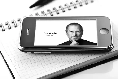 A black-and-white photo of an iPhone placed on an open notebook captures the legacy of the ultimate key man. The iPhone screen displays an image of Steve Jobs with the text "Steve Jobs 1955-2011." A black pen is positioned above the notebook in the background, adding to the poignant scene.