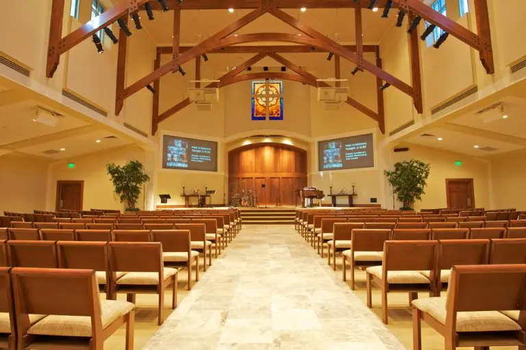 A bright, modern church interior with wooden beams and rows of cushioned pews on either side of a central aisle. The front features a stage with musical instruments, a podium, potted plants, a stained glass window, and screens displaying church information, including key man insurance for pastors.