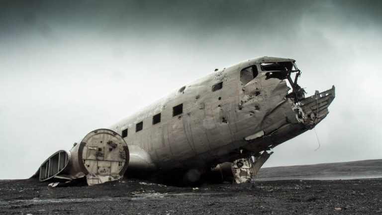 A weathered, abandoned airplane wreck lies on a desolate, rocky landscape under a cloudy, gray sky. The fuselage is noticeably damaged and deteriorated, reminiscent of the stories that underscore the need for comprehensive accidental death insurance. The scene is both eerie and forlorn.