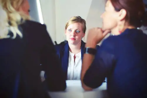 Three people are seated around a table in a meeting. The woman in the center, wearing a black blazer and white blouse, is looking intently at the two people in the foreground whose faces are blurred. Natural light filters into the room.