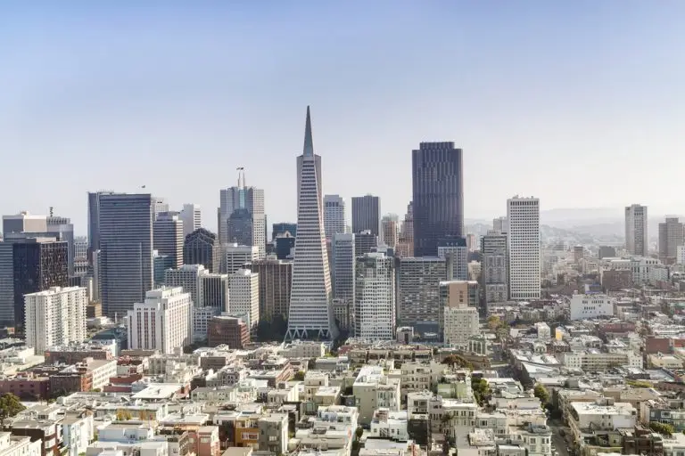 Aerial view of a cityscape featuring a mix of tall, modern skyscrapers and shorter buildings in the foreground. The tallest building, with a distinct pyramid shape, stands prominently among other high-rises under a hazy, clear sky. Residential areas and offices for key man insurance companies are visible below.