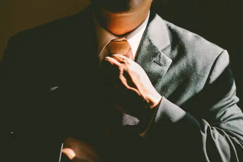 A person in a dark suit adjusts their orange necktie, possibly reflecting on executive compensation plans. The image is partially shadowed, creating a dramatic contrast between light and dark areas. Their face is not visible.