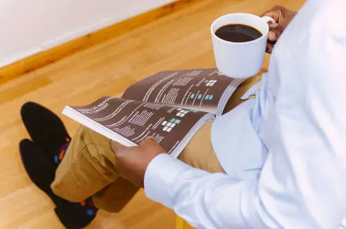 Person sitting cross-legged on the light wood floor, holding a white mug of coffee in one hand and reading a magazine about Key Man Insurance categories in the other. They are wearing a light blue shirt, beige pants, and black socks with colorful dots.