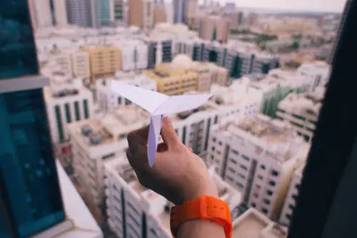 A person with an orange wristwatch is holding a white paper airplane while standing on a high floor, contemplating their Salary Continuation Plan as they overlook a bustling cityscape with numerous buildings.