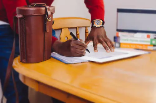 A person in a red sweater is standing at a wooden table, writing with a pen on a form likely related to structuring corporate buy-sell agreements. A brown leather bag is placed on the table, and a stack of books is visible in the background. The person is wearing a watch on their left wrist.