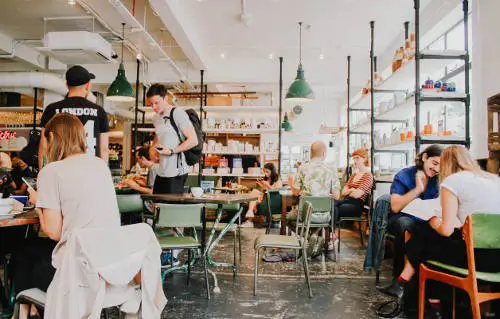 A busy cafe with people sitting at wooden tables, drinking and socializing. Some are working on laptops while others are engaged in conversations. Shelves with various items adorn the walls, and green pendant lights hang from the ceiling—a true snapshot of business owner life insurance moments.