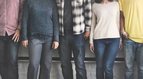 A partial view of five people standing side by side, showing only their torsos and legs, dressed in casual clothing like jeans, plaid shirts, and plain tops. They stand against a light-colored wall with a black bar at waist level, exemplifying the diverse team safeguarded by key employee insurance.