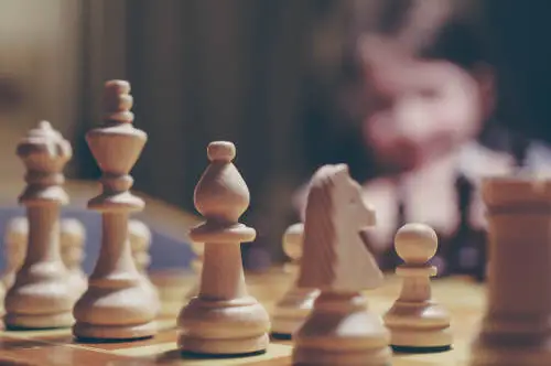 Close-up of a wooden chess set with chess pieces arranged on a board. A person is blurred in the background, focusing on the game, perhaps pondering strategic parallels to Key Man Life Insurance Alternatives. The image captures an intense and thoughtful mood.