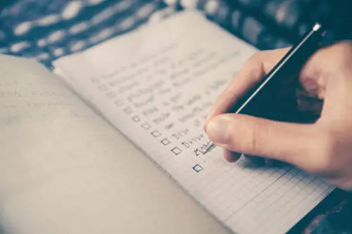 A hand holding a pen is writing on a checkered notebook. The notebook, perhaps meticulously documenting details of a supplemental executive retirement plan, contains a list with unchecked boxes next to each item. The background is soft and out of focus, emphasizing the importance of the writing task.