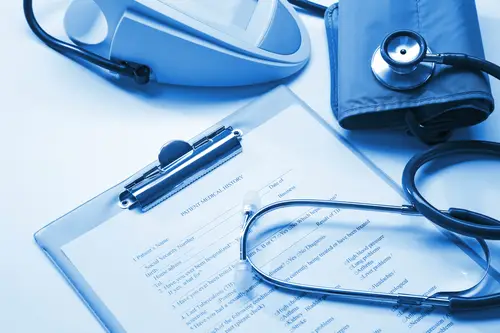 A medical-themed image featuring a stethoscope, blood pressure cuff, and a clipboard with a patient medical history form. The image has a blue tint, giving it a clinical and professional appearance, reminiscent of tools often used during a Key Man Insurance Exam.