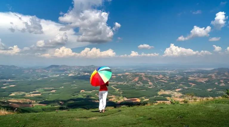 A person stands on a grassy hilltop holding a colorful umbrella, gazing at an expansive view of fields, hills, and a partly cloudy blue sky. The landscape stretches out with patchwork fields and mountainous terrain in the distance, embodying the vibrant essence of Banner Life's products.