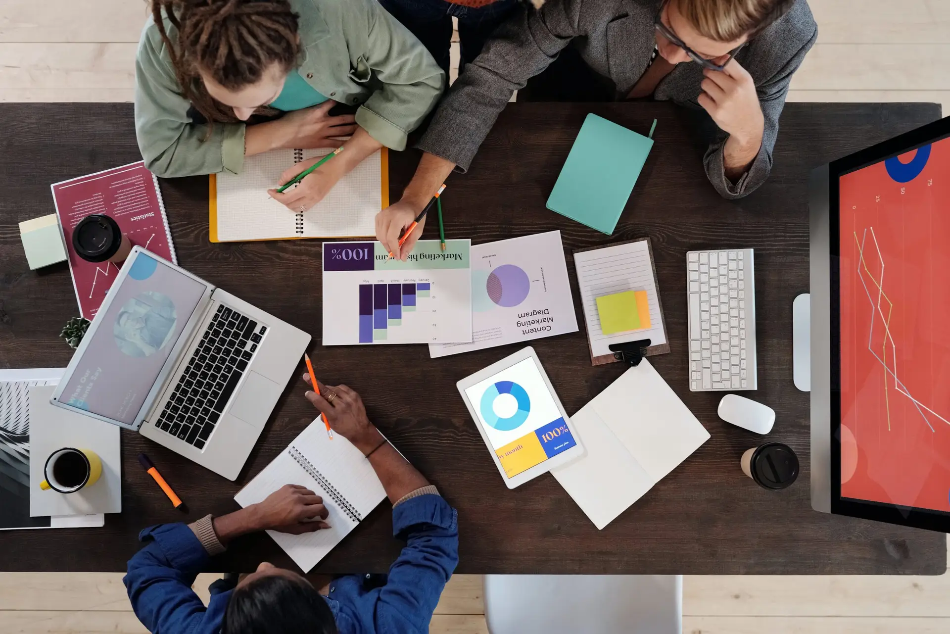 Four people are gathered around a desk covered with laptops, documents, and tablets. They appear to be in a meeting or collaborative work session, looking at charts likely related to insurance ratings and taking notes. One person is holding a pen and pointing at a document.