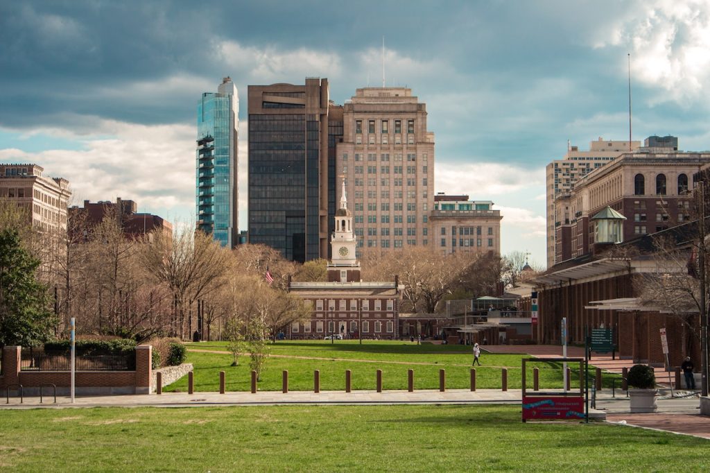 A cityscape featuring a park with green grass in the foreground. In the mid-ground, there are trees and a historic building with a clock tower. Modern skyscrapers and tall buildings dominate the background under a partly cloudy sky, symbolizing the balance of tradition and modernity similar to choosing between Whole Life or Term Policy options.