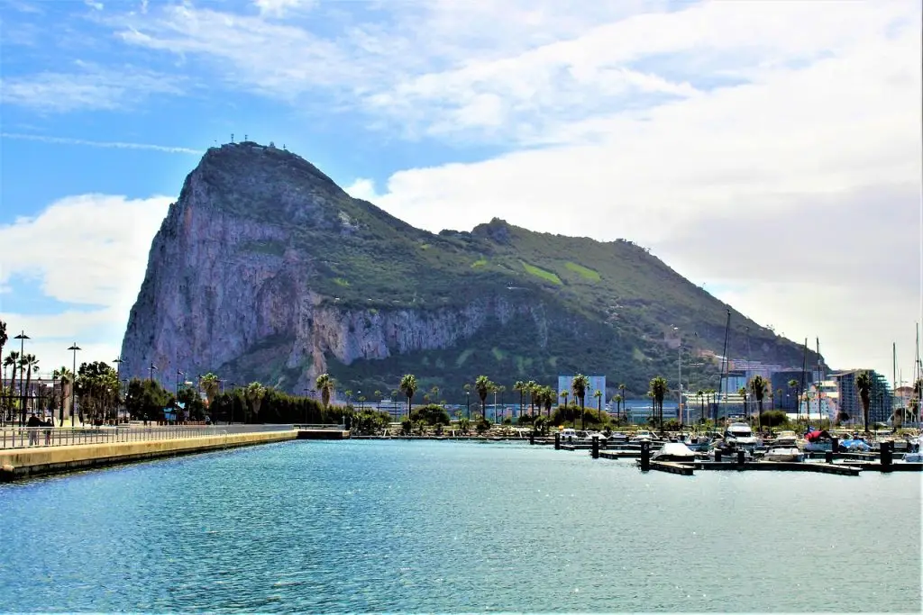 A scenic view of the Rock of Gibraltar, a prominent, steep rocky formation. Palm trees line the waterfront with a clear blue sky in the background. A marina with boats is visible along the edge of the water in the foreground, offering peace that could inspire any prudential life insurance review.