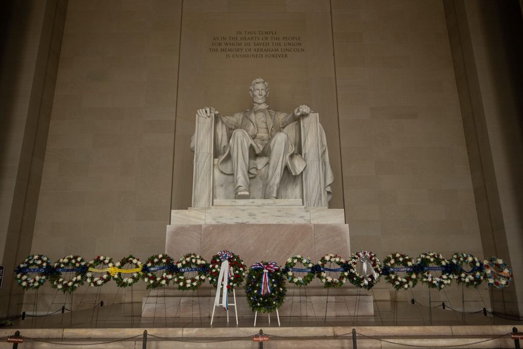 A large statue of Abraham Lincoln seated in a chair is depicted inside the Lincoln Memorial. Below the statue, numerous wreaths are placed in a row, honoring his memory. An inscription above the statue reads, "In this temple...