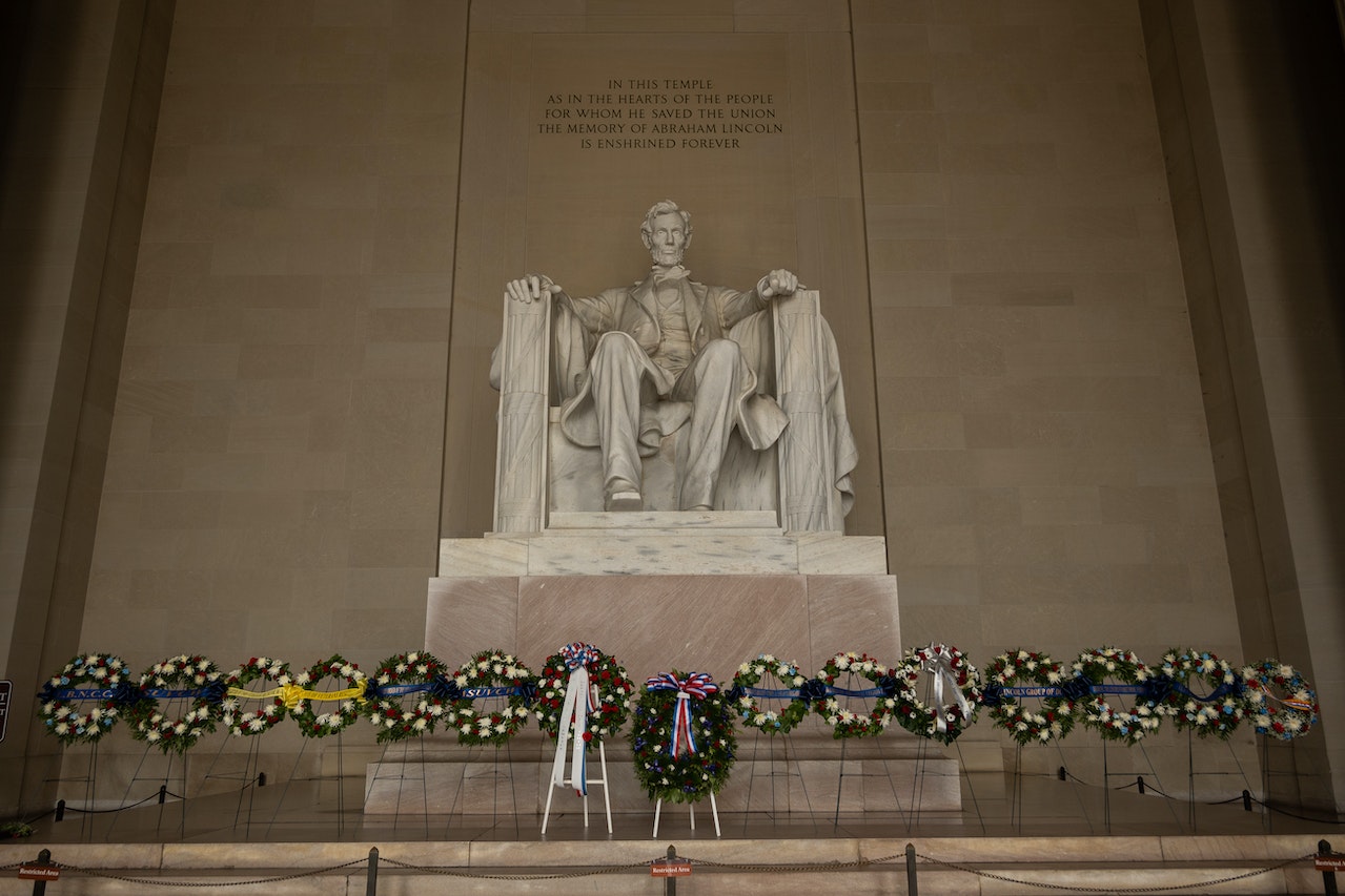 A large statue of Abraham Lincoln seated in a chair is depicted inside the Lincoln Memorial. Below the statue, numerous wreaths are placed in a row, honoring his memory. An inscription above the statue reads, "In this temple...