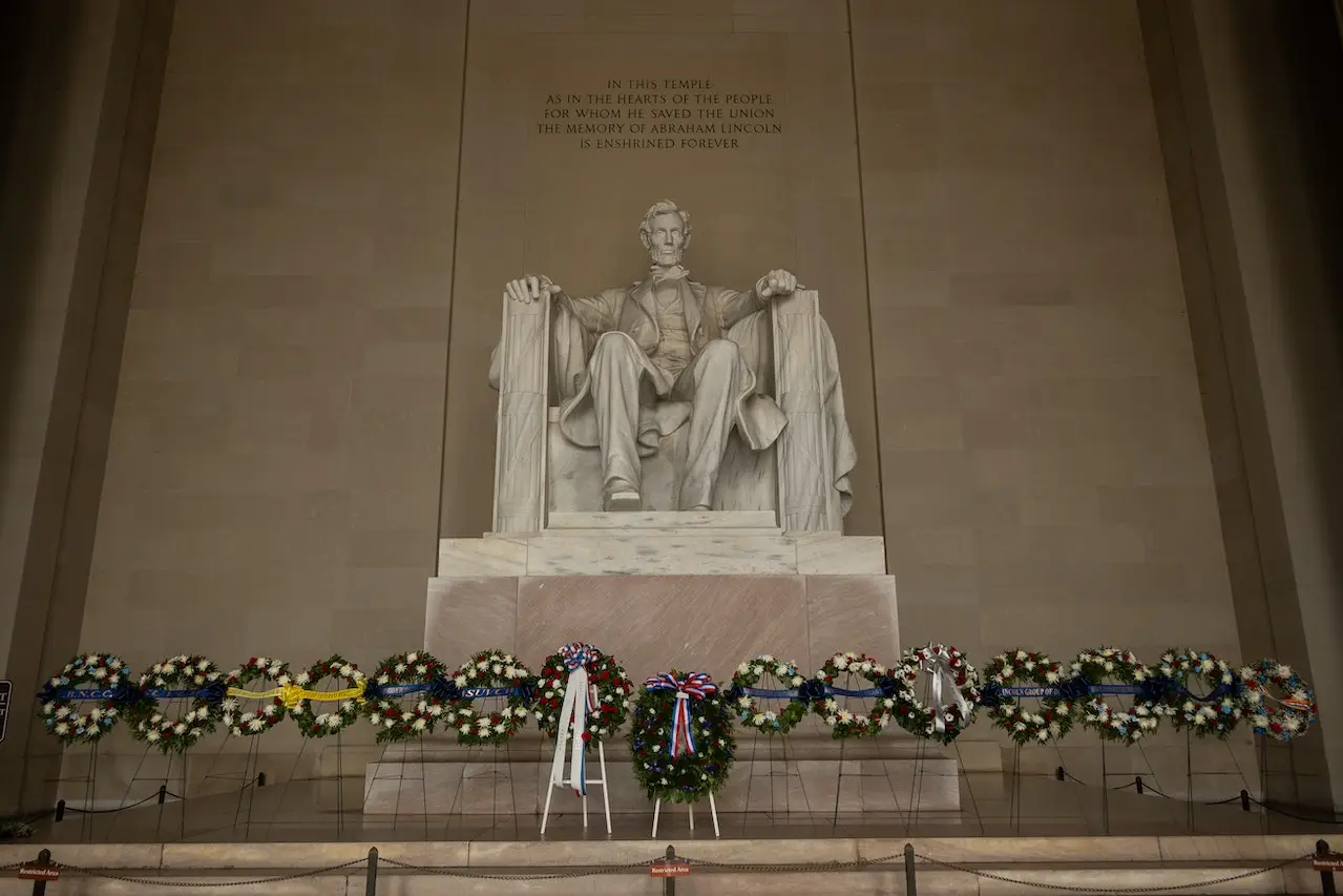 A large statue of Abraham Lincoln seated in a chair is depicted inside the Lincoln Memorial. Below the statue, numerous wreaths are placed in a row, honoring his memory. An inscription above the statue reads, "In this temple...