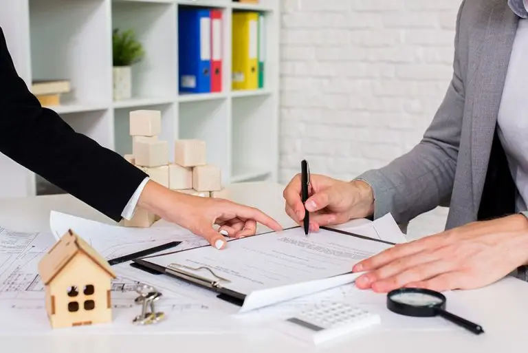 Two individuals working at a desk cluttered with documents, a small wooden house model, a magnifying glass, a calculator, and architectural plans. One is pointing at a document while the other is writing on it. Shelves with colorful folders are visible in the background.