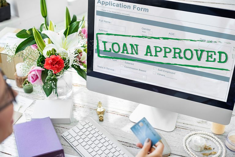 A person holding a credit card looks at a computer screen displaying an application form marked with a large "LOAN APPROVED" stamp. The desk has flowers, a purple notebook, a white keyboard, a mouse, and a gold wristwatch.