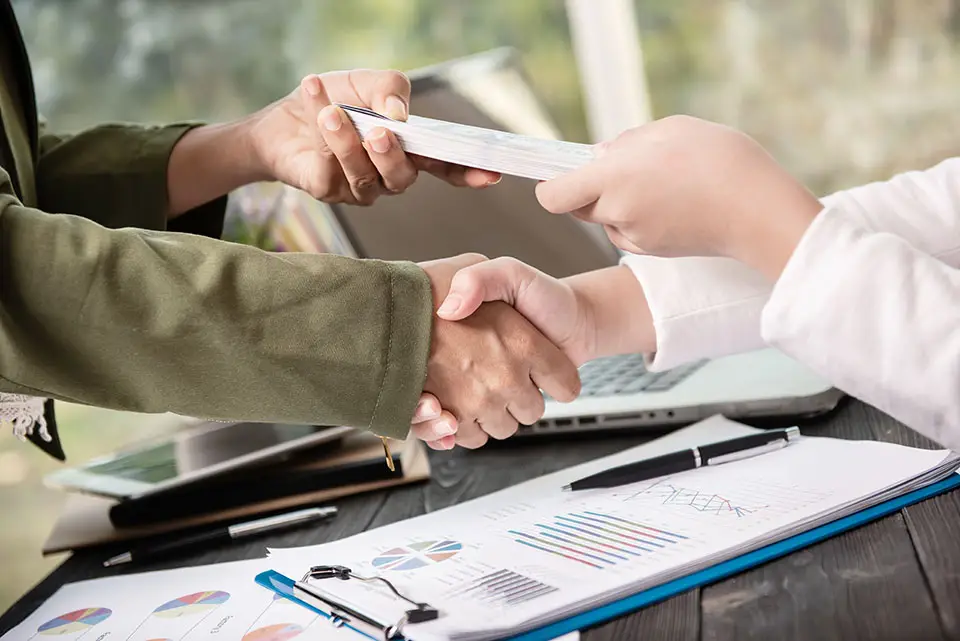 Two people shaking hands over a desk with charts, diagrams, and a laptop. One person is giving a document to the other. The setting suggests a professional business environment, possibly indicating the closing of a deal or agreement.