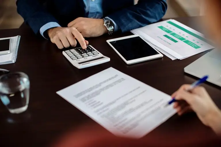 Two people are seated at a desk with documents, a calculator, a tablet, and a glass of water. One person is using the calculator, pointing at it, while the other is holding a pen and writing on a piece of paper. An open notebook and papers are also visible.